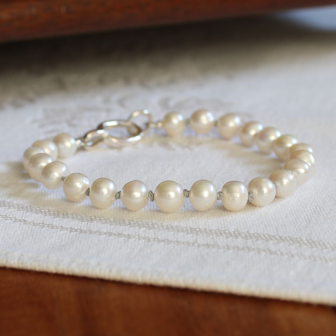 A strand bracelet made of round white pearls hand-knotted with silver silk and a silver clasp, resting on a white textured fabric surface.