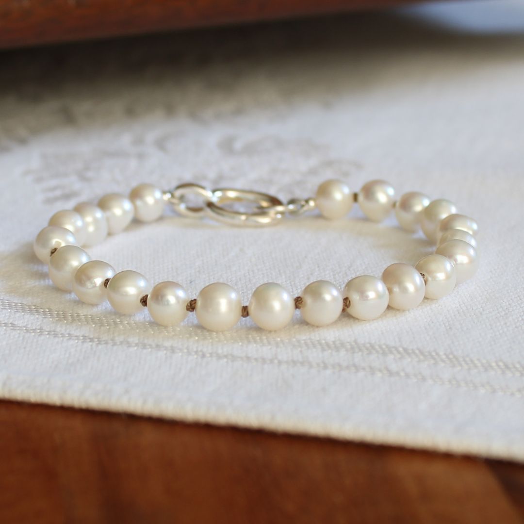 A strand bracelet made of round white pearls hand-knotted smoke silk , with a sterling silver clasp, resting on a white textured fabric over a wooden surface.