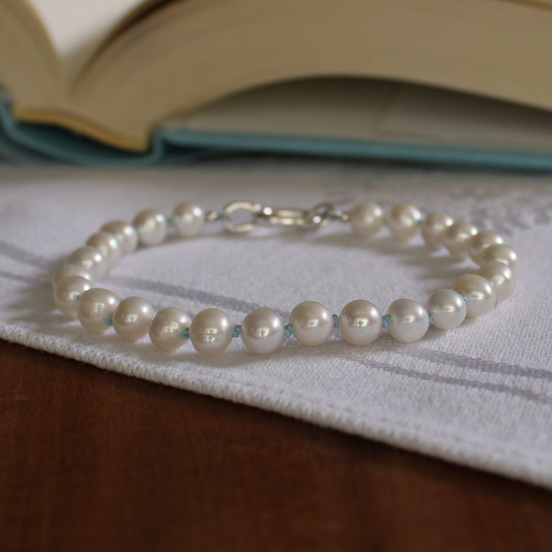 A single-strand pearl bracelet hand-knotted with turquoise silk, with a silver clasp resting on a white textured cloth over a wooden surface, with an open book blurred in the background.