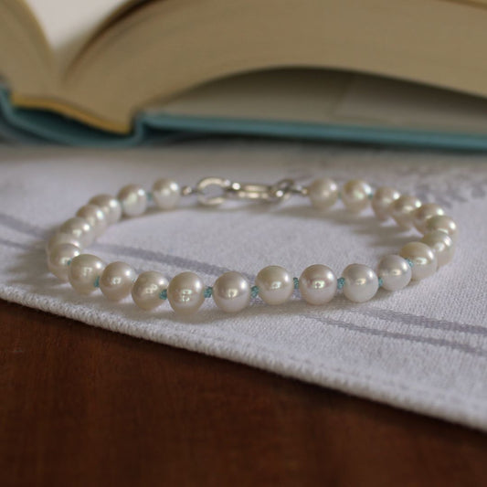 A single-strand pearl bracelet hand-knotted with turquoise silk, with a silver clasp resting on a white textured cloth over a wooden surface, with an open book blurred in the background.