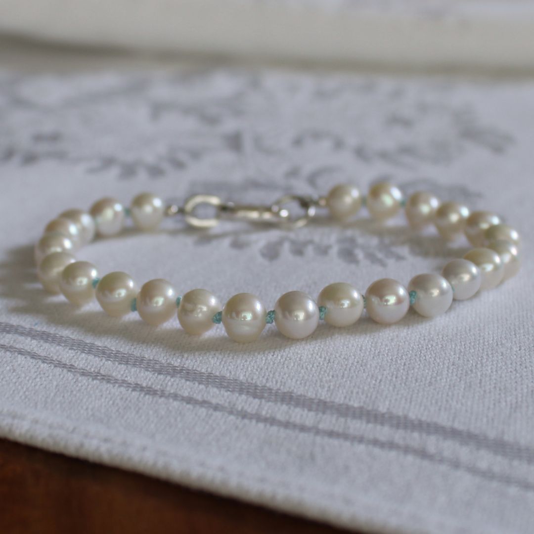 A pearl beaded bracelet, knotted with turquoise silk,  with a silver clasp resting on a white embroidered fabric surface.
