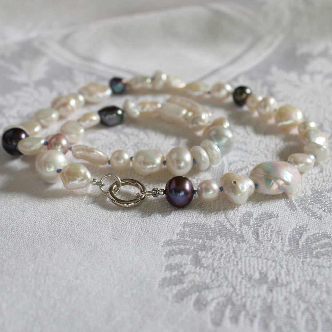 One-of-a-kind mixed pearl necklace with keshi, coin, round, baroque, and peacock pearls hand-knotted on delft silk, and showing 13mm sterling silver round charmer clasp, laying on a linen cloth.