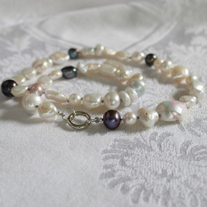 One-of-a-kind mixed pearl necklace with keshi, coin, round, baroque, and peacock pearls hand-knotted on delft silk, and showing 13mm sterling silver round charmer clasp, laying on a linen cloth.