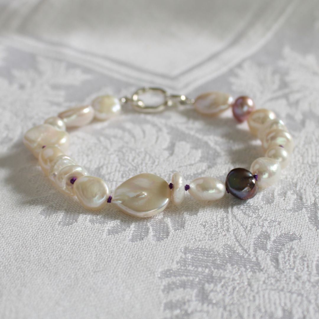A mixed pearl bracelet featuring coin, keshi, peacock, and baroque pearls hand-knotted on amethyst silk displayed on a textured white fabric background.