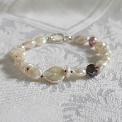 A mixed pearl bracelet featuring coin, keshi, peacock, and baroque pearls hand-knotted on amethyst silk displayed on a textured white fabric background.
