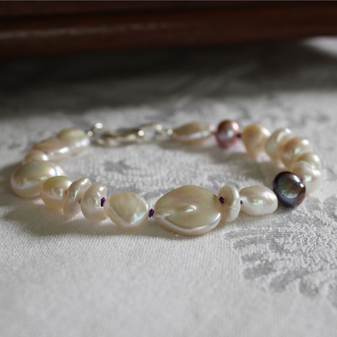 A mixed bracelet made of white baroque, coin, keshi, and peacock pearls hand-knotted on amethyst silk, displayed on a textured white fabric surface.