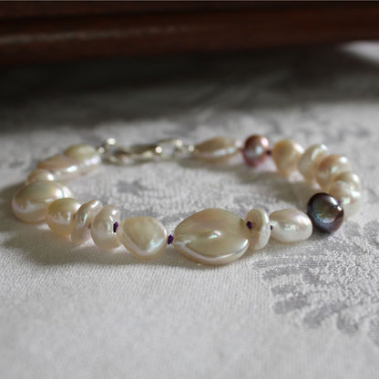 A mixed bracelet made of white baroque, coin, keshi, and peacock pearls hand-knotted on amethyst silk, displayed on a textured white fabric surface.