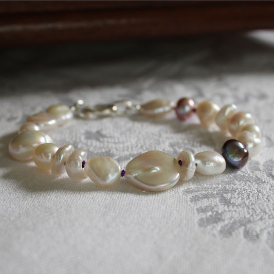A mixed bracelet made of white baroque, coin, keshi, and peacock pearls hand-knotted on amethyst silk, displayed on a textured white fabric surface.
