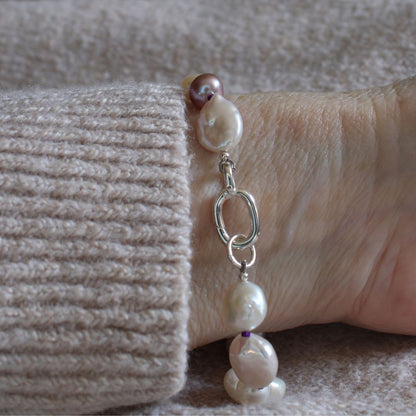 A close up of a wrist wearing a mixed pearl bracelet of coin, keshi, peacock, and baroque pearls hand-knotted on amethyst silk, showing the sterling silver easy close clasp.
