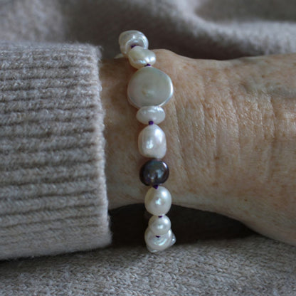 A close-up of a mixed pearl bracelet worn on a wrist, featuring coin, keshi, baroque, and peacock pearls hand-knotted on amethyst silk, styled with a beige ribbed knit cuff against a soft beige background.