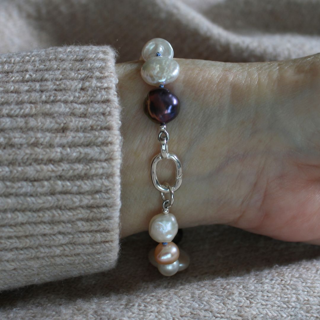 A close-up of a person's wrist wearing a mixed pearl bracelet. The bracelet features a mix of white, cream, and dark purple pearls with a sterling silver easy close clasp.