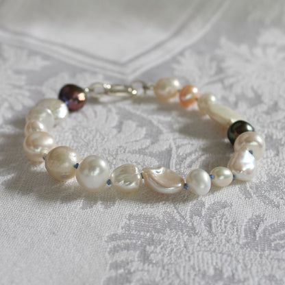 A bracelet made of mixed pearls including peacock, keshi, and baroque pearls hand-knotted on delft silk, resting on a white patterned fabric surface.