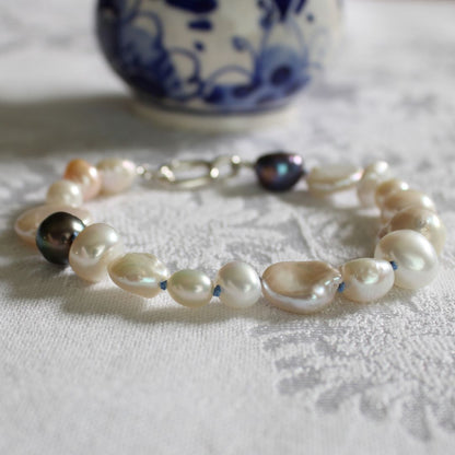 A bracelet made of mixed white, cream and peacock pearls hand-knotted on delft silk, displayed on a white textured fabric surface with a blue-and-white ceramic vase blurred in the background.