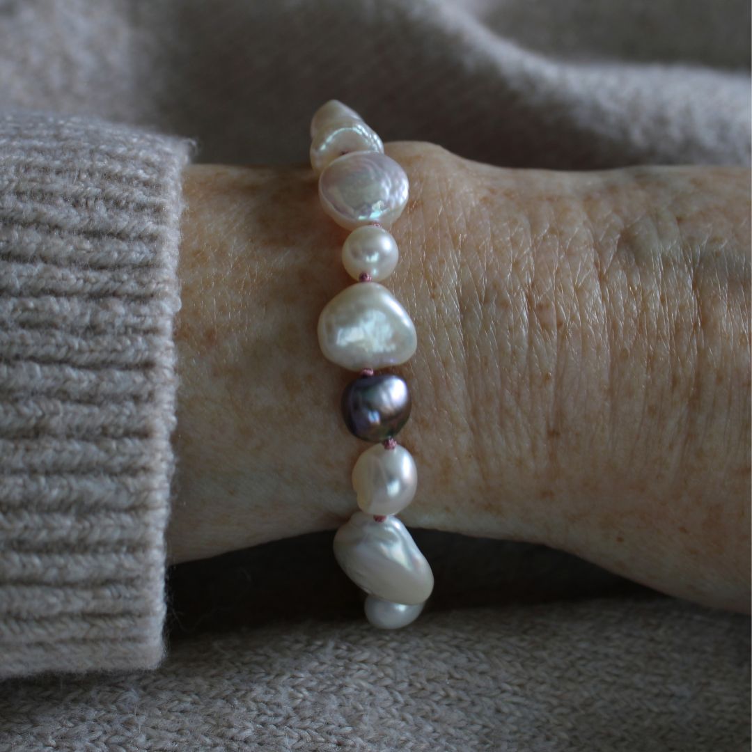 A close-up of a person's wrist wearing a mixed pearl bracelet made of keshi, round, baroque, peacock pearls hand-knotted on old rose silk, styled with a beige knit sweater cuff.