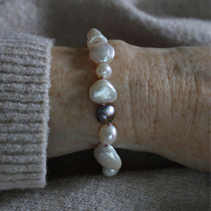 A close-up of a person's wrist wearing a mixed pearl bracelet made of keshi, round, baroque, peacock pearls hand-knotted on old rose silk, styled with a beige knit sweater cuff.