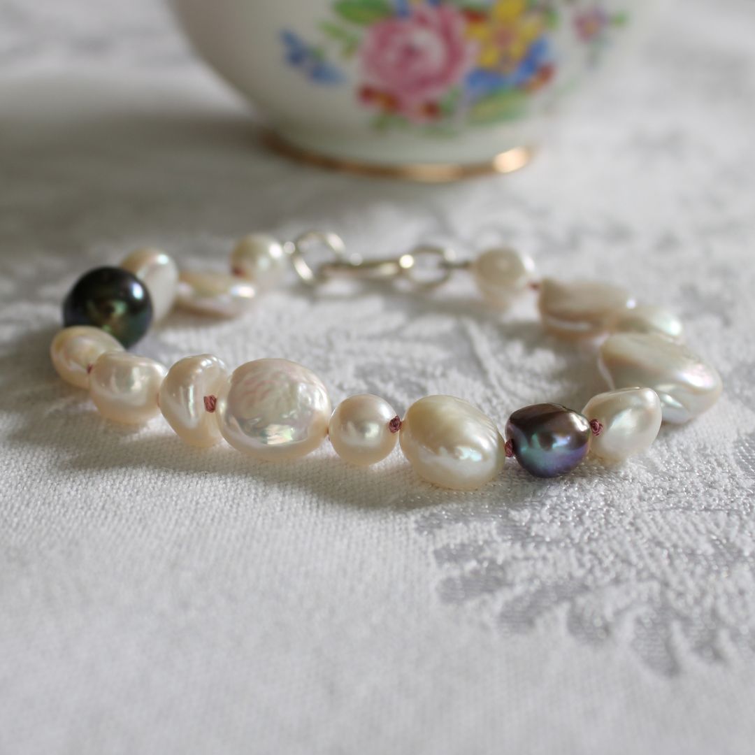 A mixed pearl bracelet made of keshi, round, baroque, peacock pearls hand-knotted on old rose silk, resting on a white textured fabric surface with a blurred floral ceramic bowl in the background.