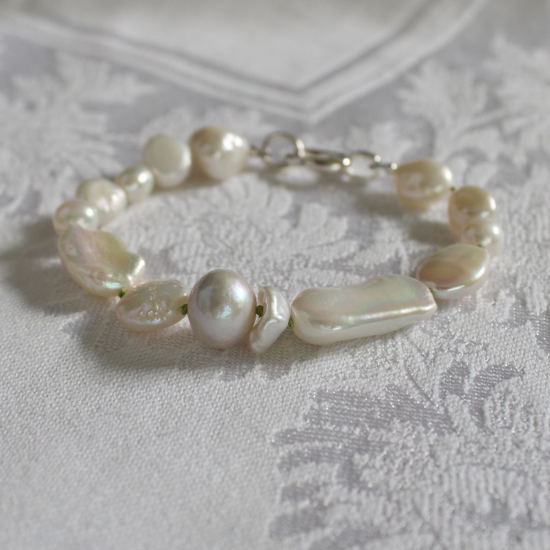 A mixed pearl bracelet hand-knotted on peridot silk with a silver clasp, resting on a white textured lace fabric background.