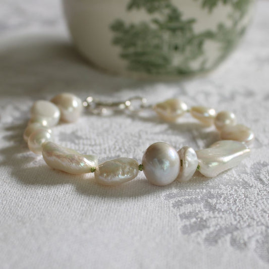 A mixed pearl bracelet hand-knotted with peridot silk with a silver clasp, resting on a white textured lace fabric with a blurred green and white ceramic cup in the background.