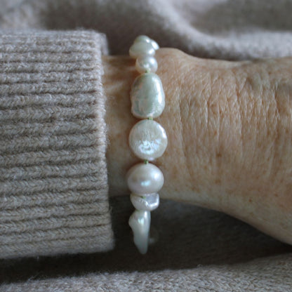 A close-up of a person's wrist wearing a bracelet made of irregular white baroque pearls hand-knotted on peridot silk, paired with a beige ribbed knit sweater cuff.