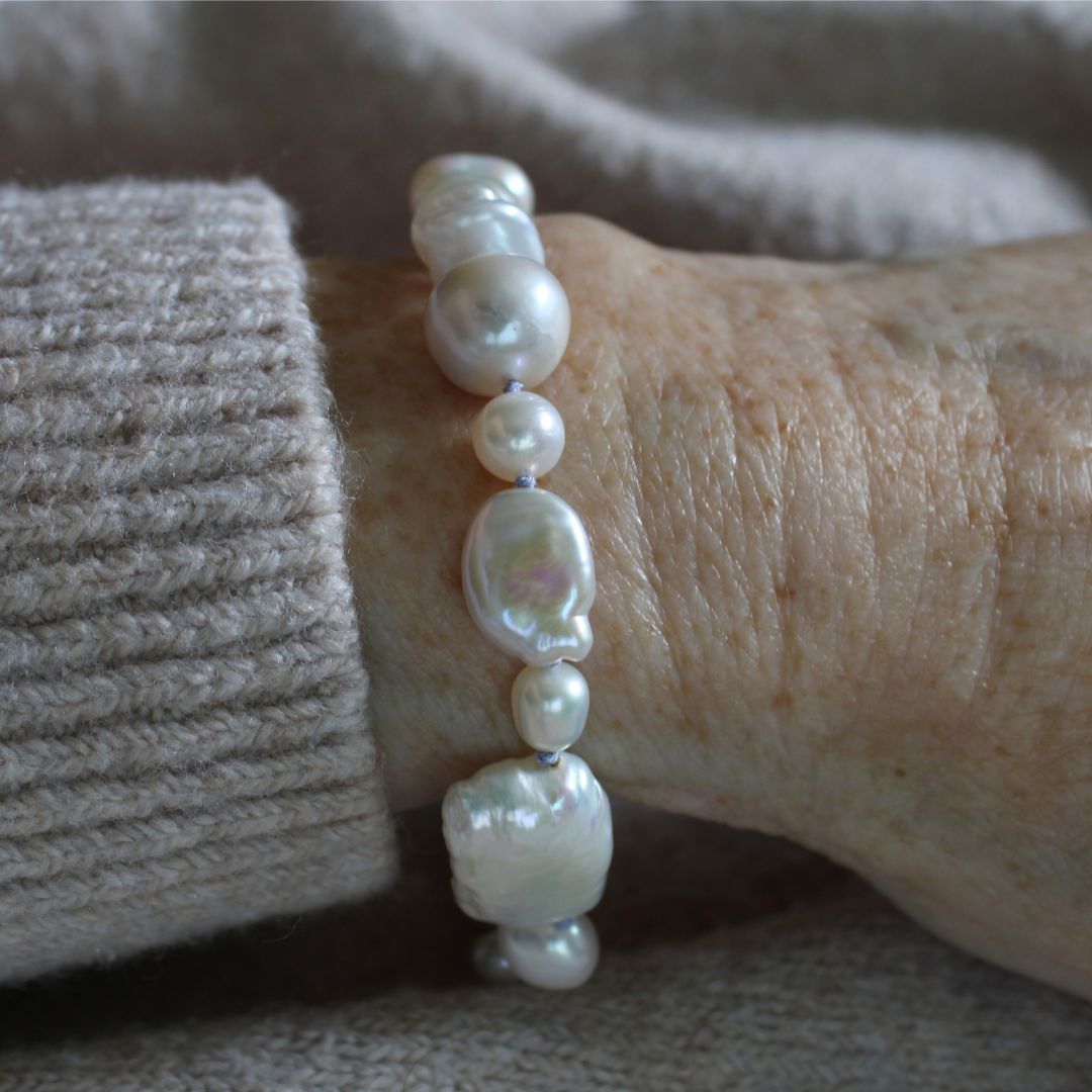 A close-up of a person's wrist wearing a mixed pear bracelet hand-knotted on periwinkle silk, paired with a ribbed beige knit cuff.