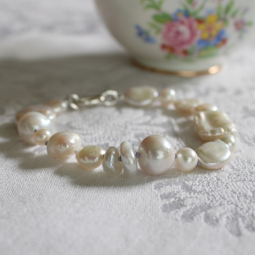 A mixed pearl bracelet hand-knotted on periwinkle silk, with a silver clasp, resting on a white textured fabric surface with a blurred floral ceramic bowl in the background.