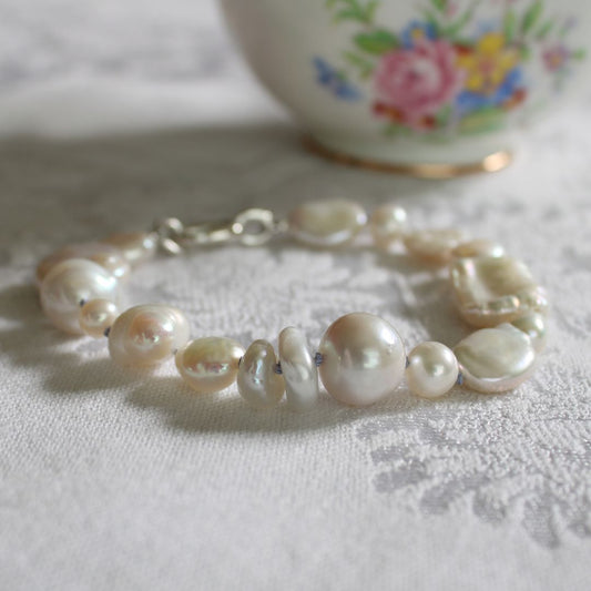A mixed pearl bracelet hand-knotted on periwinkle silk, with a silver clasp, resting on a white textured fabric surface with a blurred floral ceramic bowl in the background.