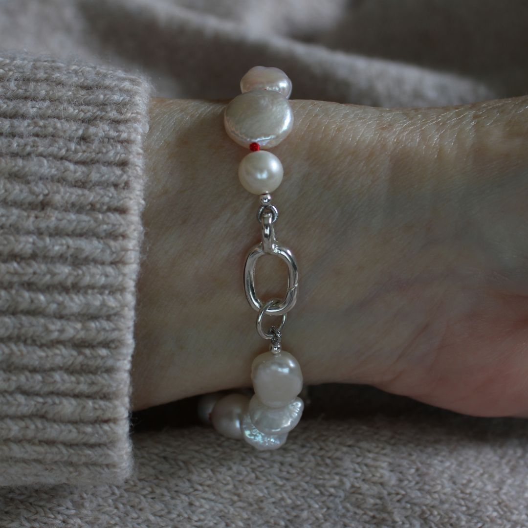 A close-up of a mixed pearl bracelet hand-knotted on ruby silk and showing the sterling silver easy close clasp, worn on a wrist covered by a beige ribbed knit sleeve.