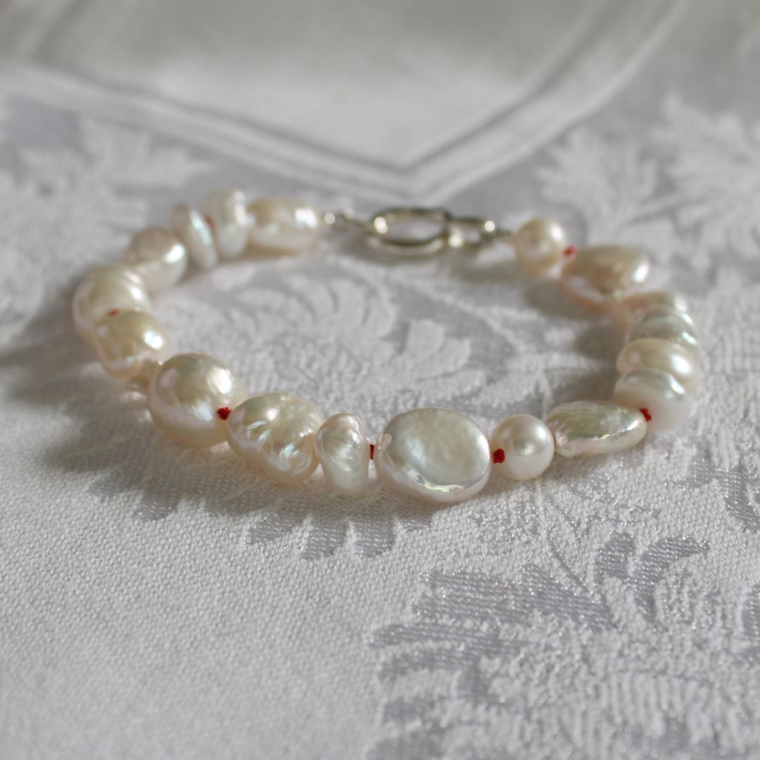 A mixed pearl bracelet hand-knotted with ruby silk and a silver clasp, resting on a white patterned fabric surface.