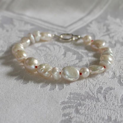 A mixed pearl bracelet hand-knotted with ruby silk and a silver clasp, resting on a white patterned fabric surface.