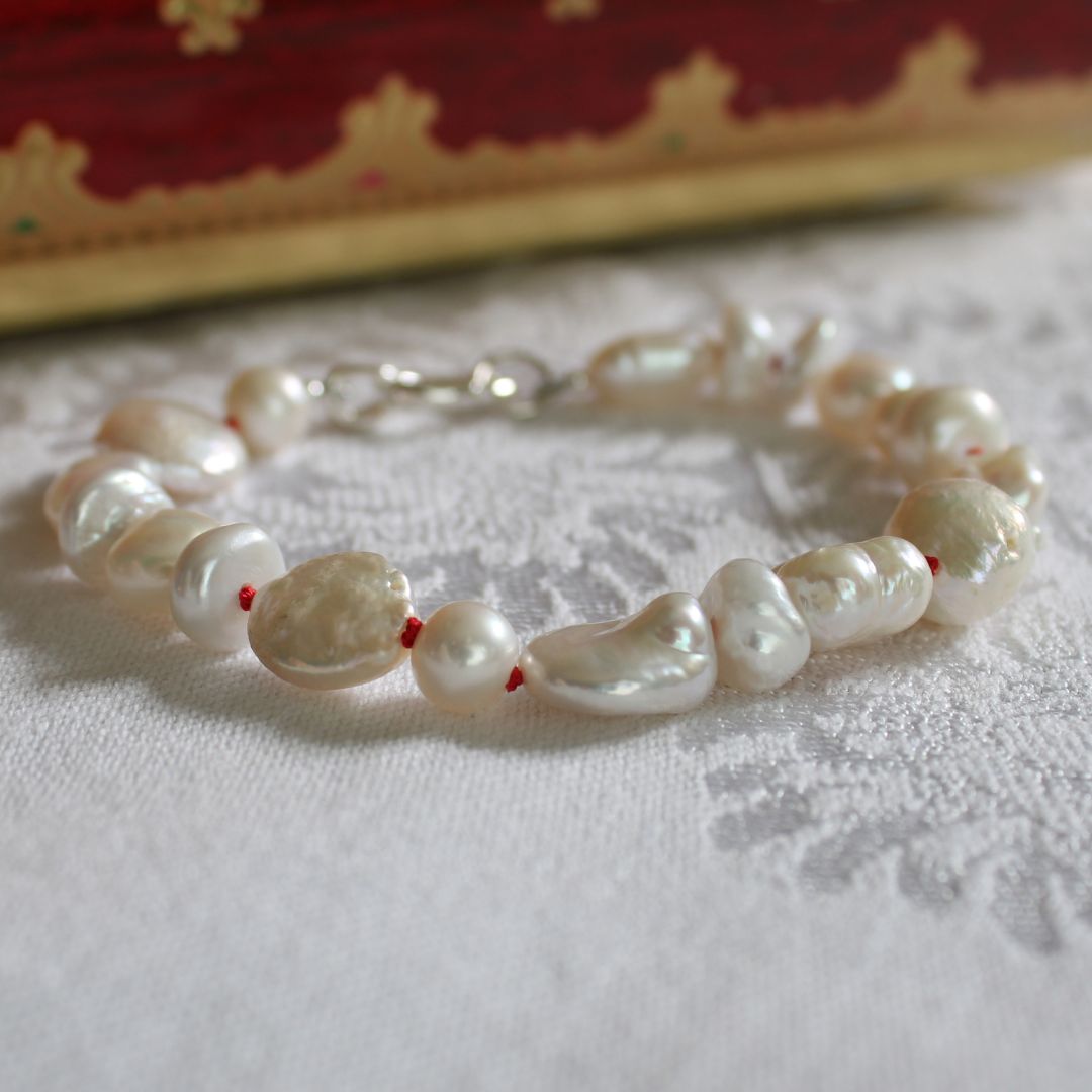A mixed pearl bracelet hand-knotted on ruby silk, resting on a white textured fabric surface with a blurred red and gold box in the background.