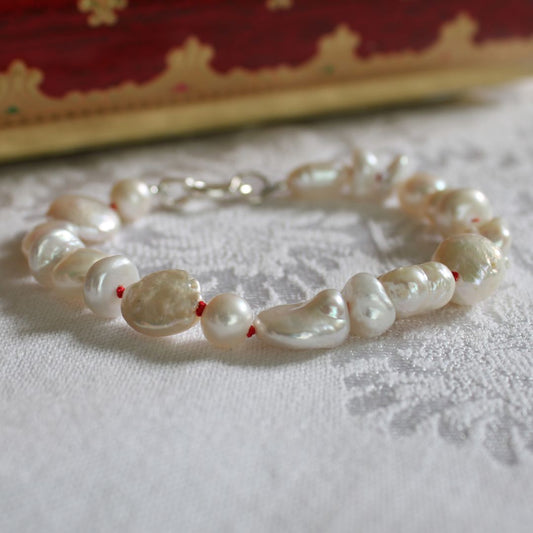 A mixed pearl bracelet hand-knotted on ruby silk, resting on a white textured fabric surface with a blurred red and gold box in the background.