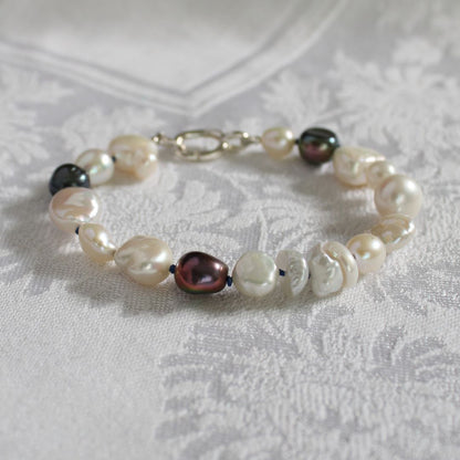A mixed pearl bracelet made of keshi, round, baroque, peacock pearls hand-knotted on sapphire silk, resting on a white damask fabric background.