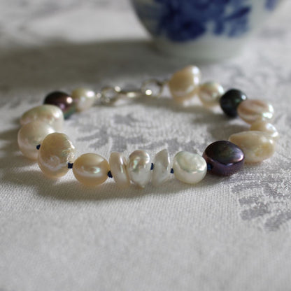 A mixed pearl bracelet made of keshi, round, baroque, peacock pearls hand-knotted on sapphire silk, resting on a linen cloth with blue and white cup in background. 