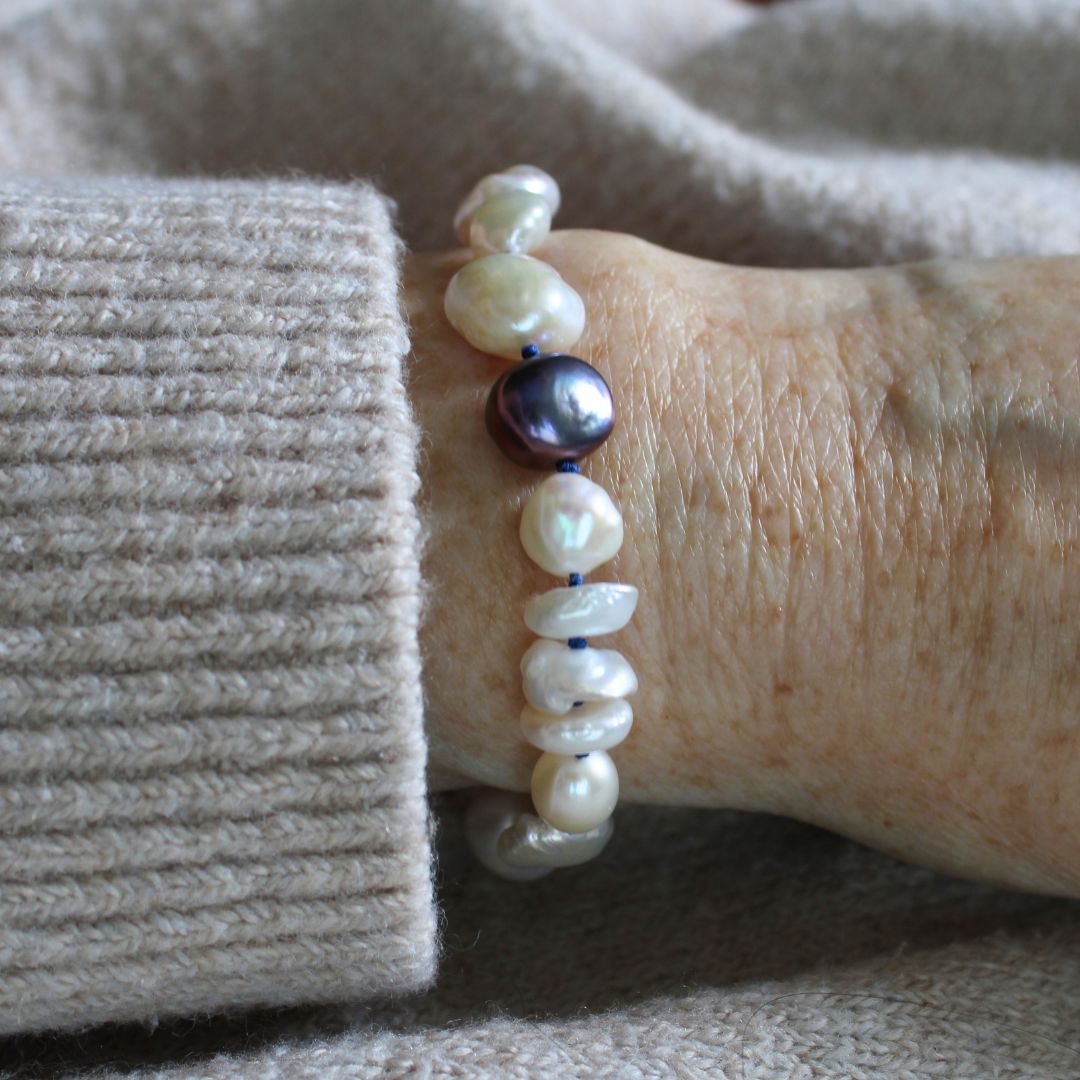 A close-up of a person's wrist wearing a mixed pearl bracelet made of keshi, round, baroque, peacock pearls hand-knotted on sapphire silk, set against a beige knit sweater cuff and soft neutral fabric background.