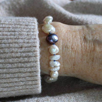 A close-up of a person's wrist wearing a mixed pearl bracelet made of keshi, round, baroque, peacock pearls hand-knotted on sapphire silk, set against a beige knit sweater cuff and soft neutral fabric background.