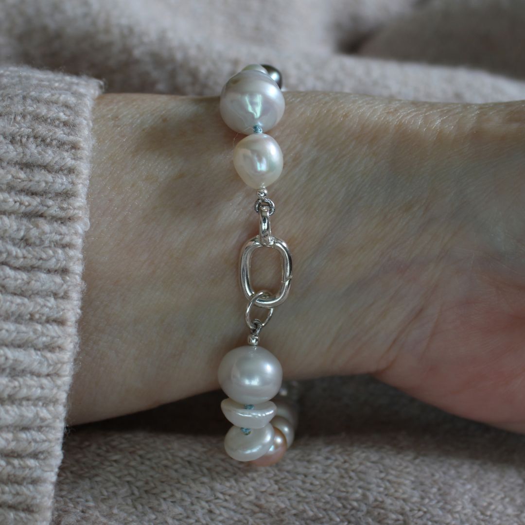A close-up of a person's wrist wearing A mixed pearl bracelet made of keshi, round, baroque, peacock pearls hand-knotted on turquoise silk, showing the sterling silver easy to close clasp, set against a soft beige knit sweater.