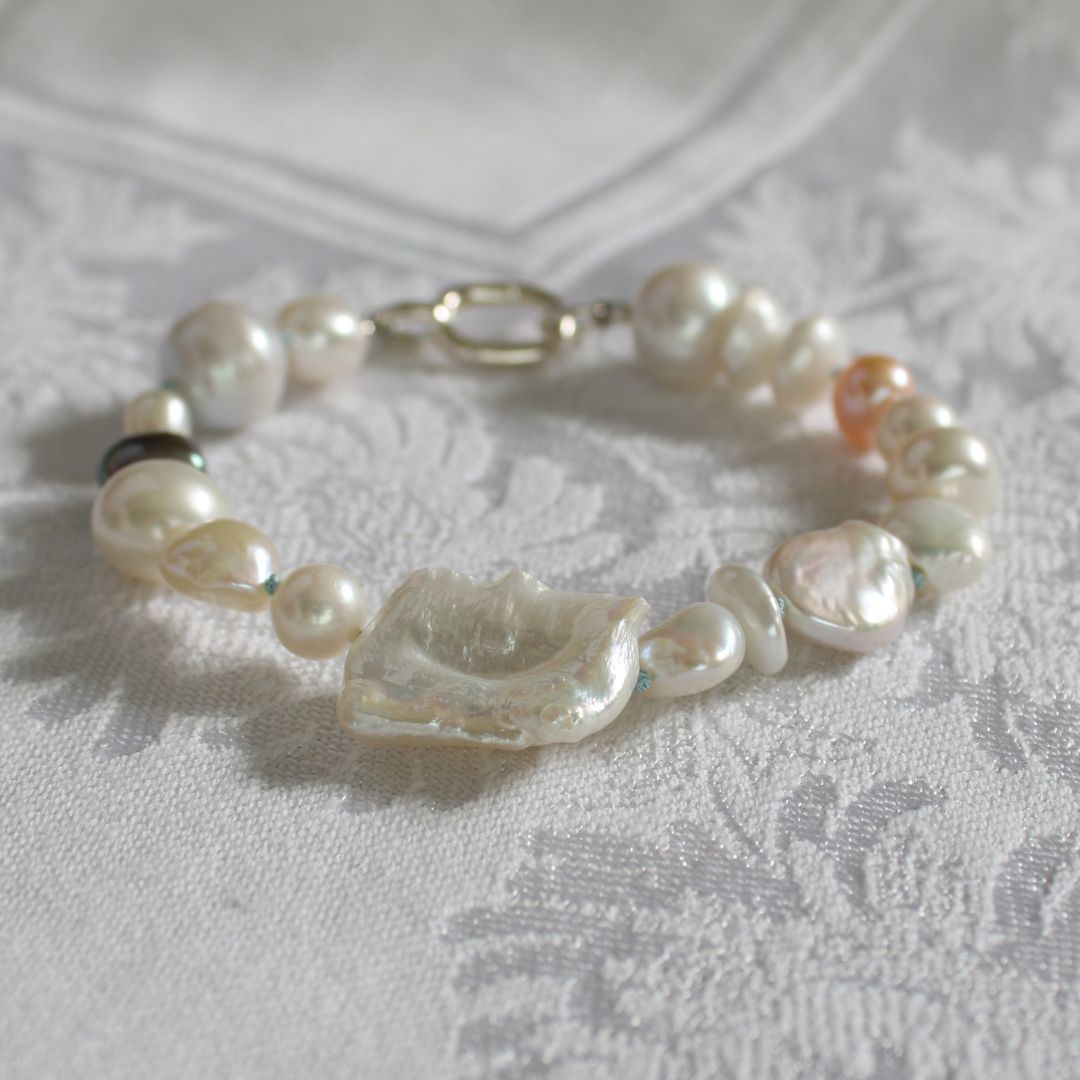 A mixed pearl bracelet made of keshi, round, baroque, peacock pearls hand-knotted on turquoise silk, resting on a white textured fabric background.