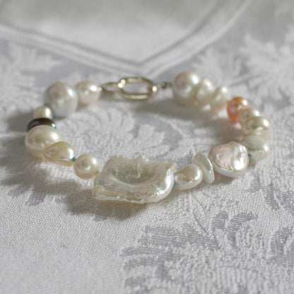 A mixed pearl bracelet made of keshi, round, baroque, peacock pearls hand-knotted on turquoise silk, resting on a white textured fabric background.