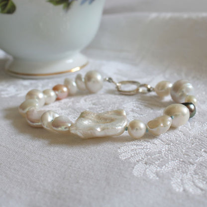 A mixed pearl bracelet made of keshi, round, baroque, peacock pearls hand-knotted on turquoise silk, resting on a linen cloth with pale turquoise cup in background.