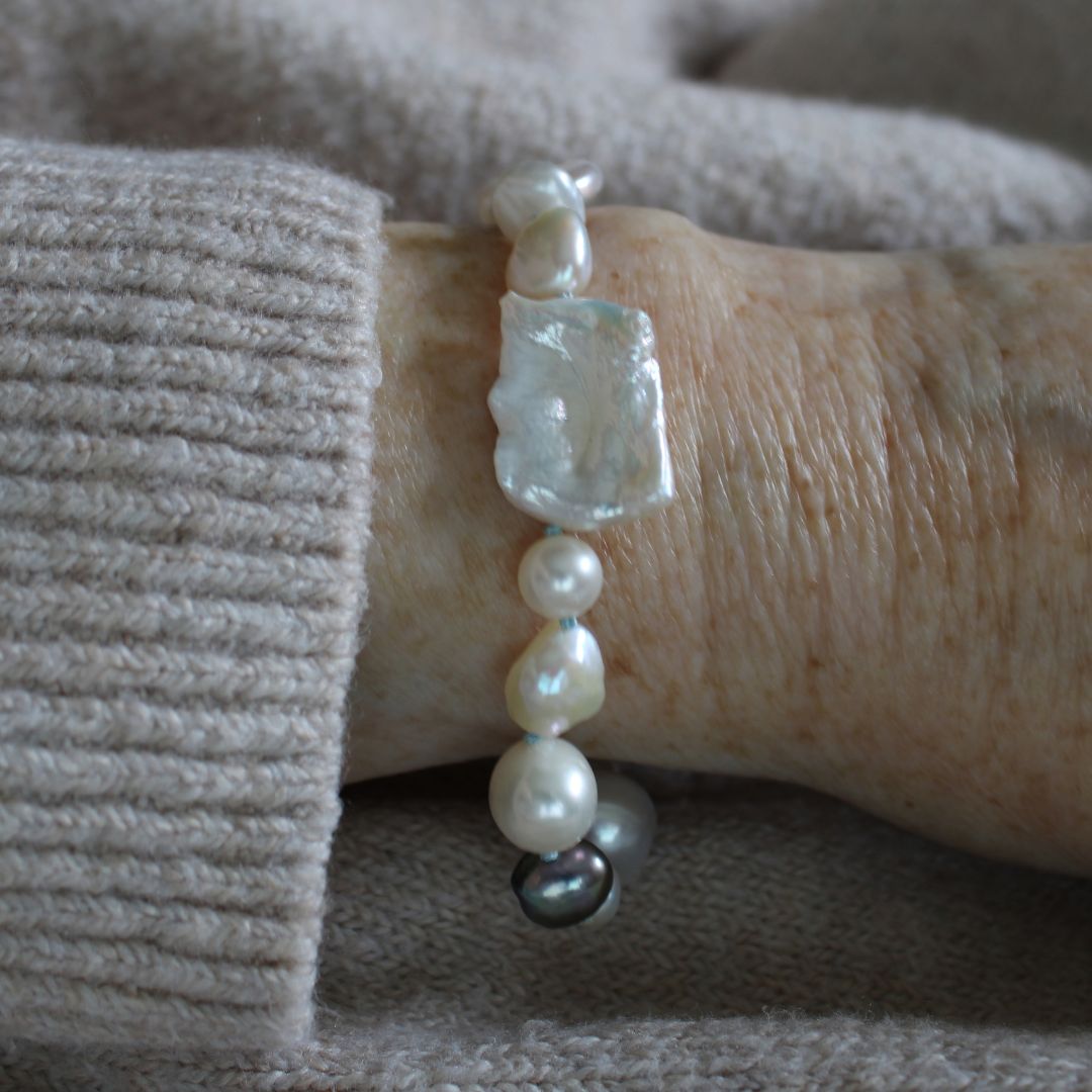 A close-up of a mixed pearl bracelet made of keshi, round, baroque, peacock pearls hand-knotted on turquoise silk, on a wrist, set against a beige knit sweater.