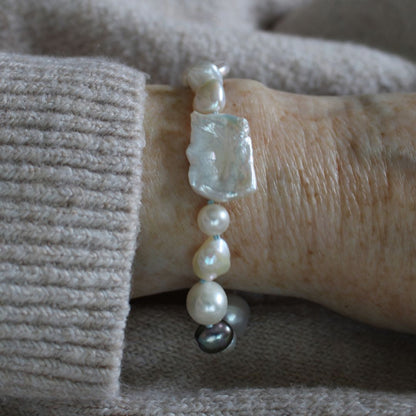 A close-up of a mixed pearl bracelet made of keshi, round, baroque, peacock pearls hand-knotted on turquoise silk, on a wrist, set against a beige knit sweater.