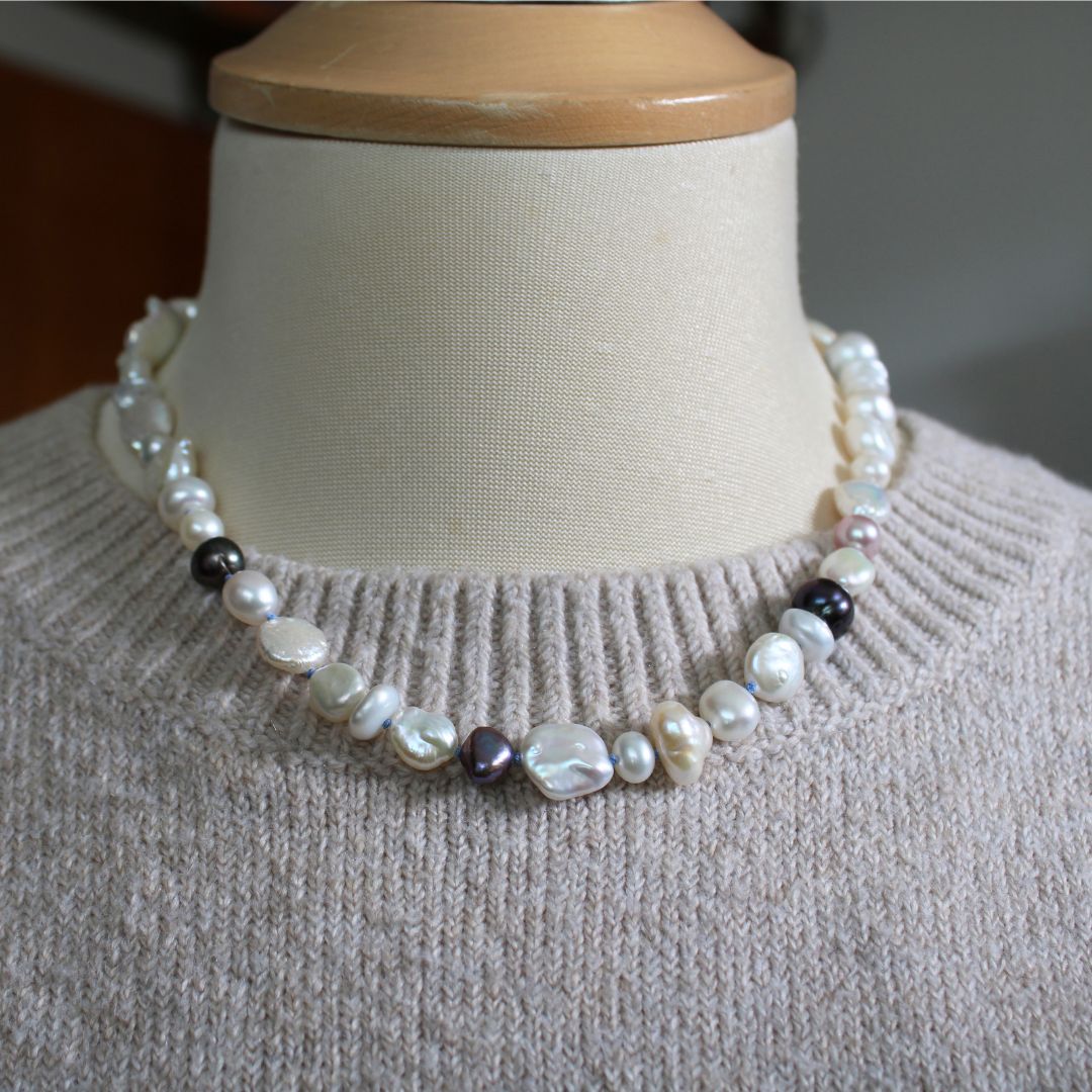 One-of-a-kind mixed pearl necklace with keshi, coin, round, baroque, and peacock pearls hand-knotted on delft silk, shown on mannequin with beige knitted sweater.