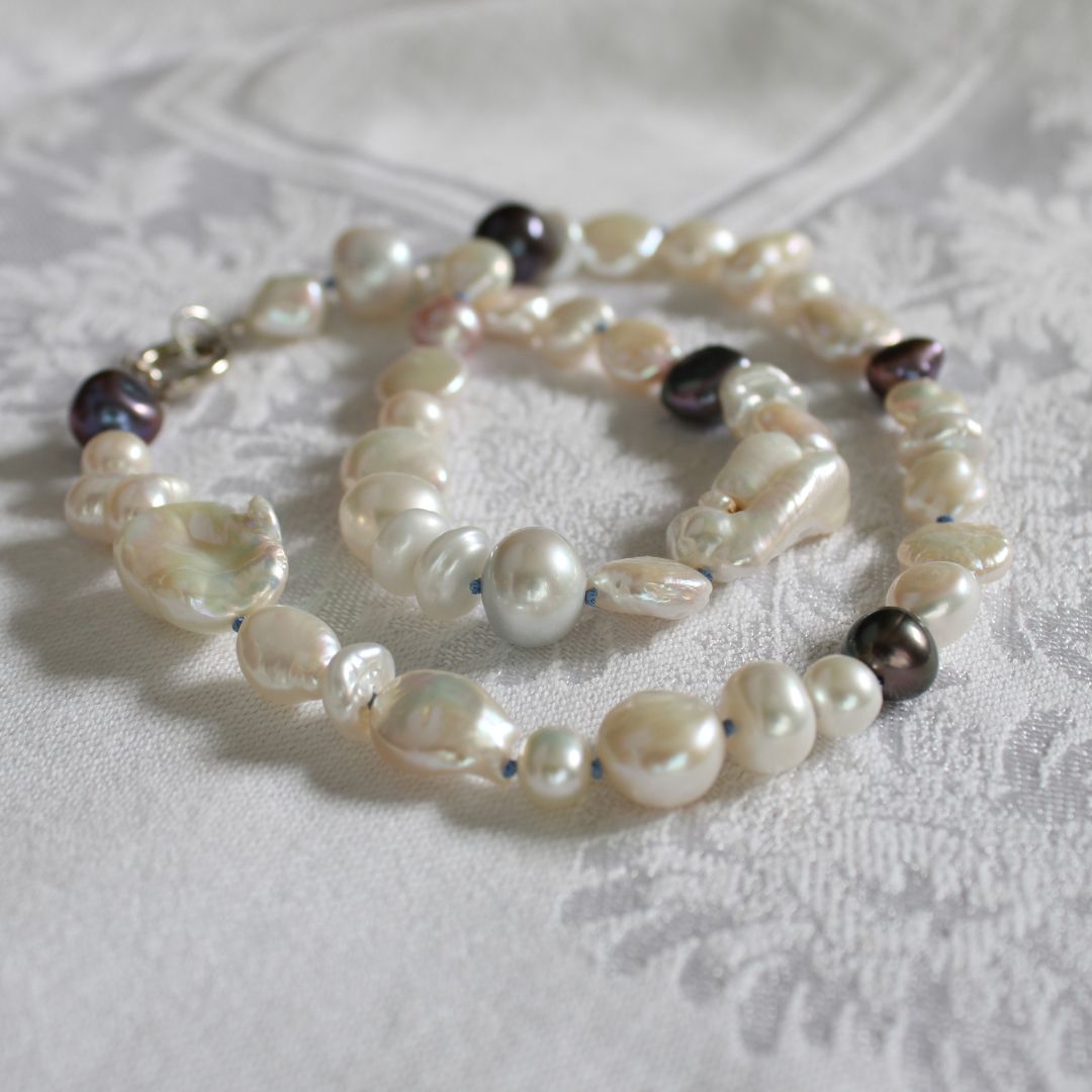 One-of-a-kind mixed pearl necklace with keshi, coin, round, baroque, and peacock pearls hand-knotted on delft silk, laying on a linen cloth.
