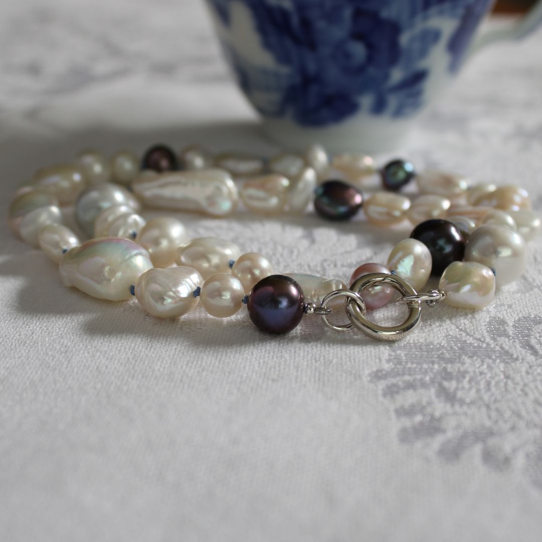 One-of-a-kind mixed pearl necklace with keshi, coin, round, baroque, and peacock pearls hand-knotted on delft silk, and showing 13mm sterling silver round charmer clasp, laying on a linen cloth with blue and with cup in background. 