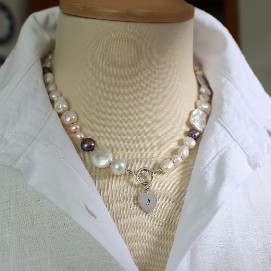One-of-a-kind mixed pearl necklace with keshi, coin, round, baroque, and peacock pearls hand-knotted on ruby silk, and showing 13mm sterling silver round charmer clasp and hand-stamped initial heart charm, shown on mannequin with white linen blouse with upturned collar.