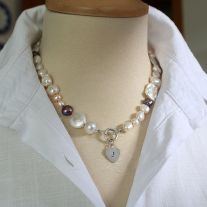 One-of-a-kind mixed pearl necklace with keshi, coin, round, baroque, and peacock pearls hand-knotted on ruby silk, and showing 13mm sterling silver round charmer clasp and hand-stamped initial heart charm, shown on mannequin with white linen blouse with upturned collar.