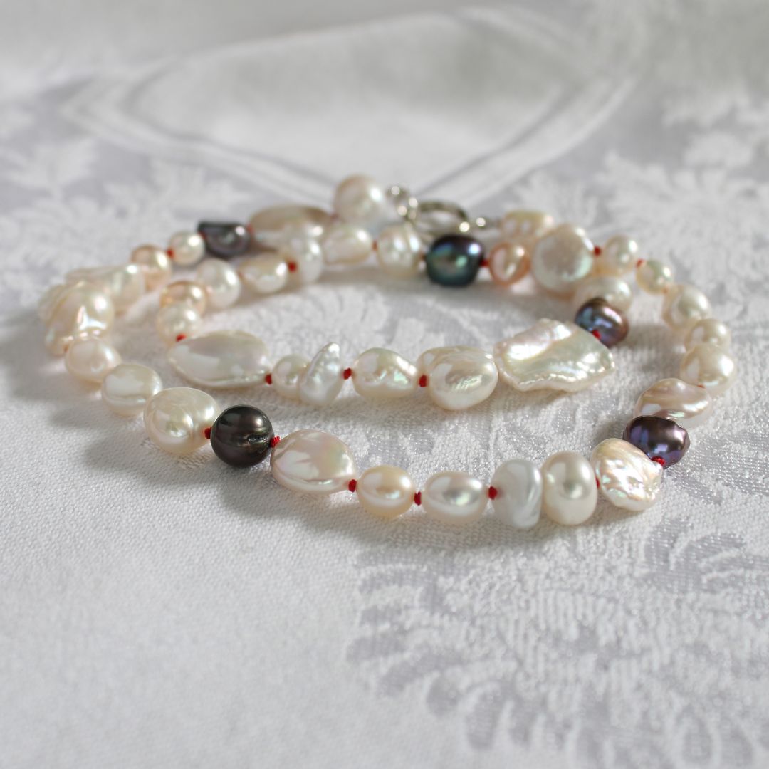 One-of-a-kind mixed pearl necklace with keshi, coin, round, baroque, and peacock pearls hand-knotted on ruby silk, laying on a linen cloth. 