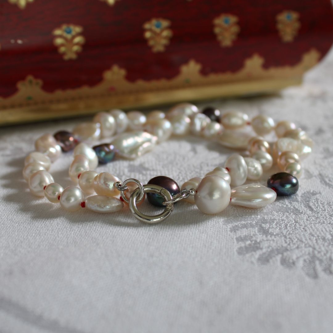 One-of-a-kind mixed pearl necklace with keshi, coin, round, baroque, and peacock pearls hand-knotted on ruby silk, and showing 13mm sterling silver round charmer clasp, laying on a linen cloth. 